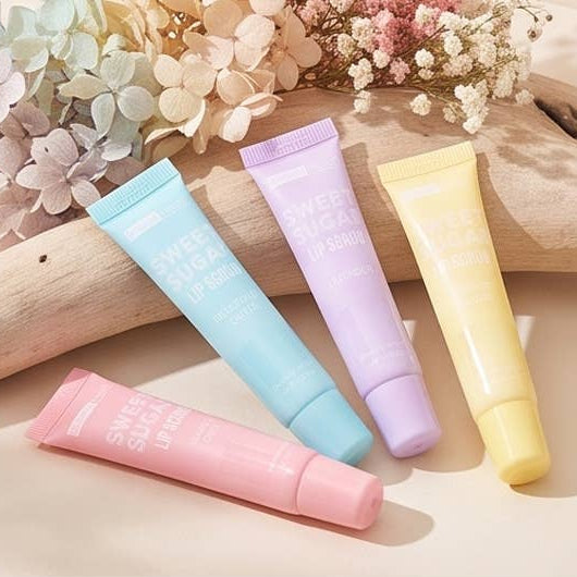 Four lip serum tubes in pastel pink, light blue, lavender, and pale yellow, arranged on a beige surface with dried flowers and a wooden prop in the background.