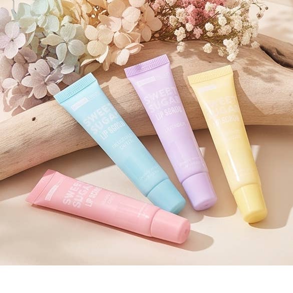 Four lip serum tubes in pastel pink, light blue, lavender, and pale yellow, arranged on a beige surface with dried flowers and a wooden prop in the background.