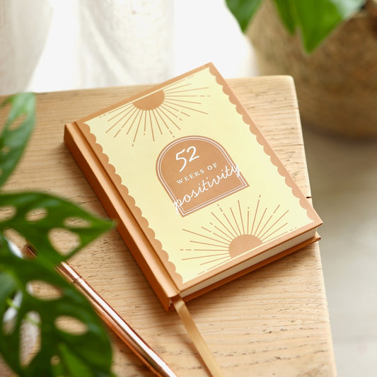 Notebook with '52 Weeks of Positivity' on a wooden surface with plants in the background
