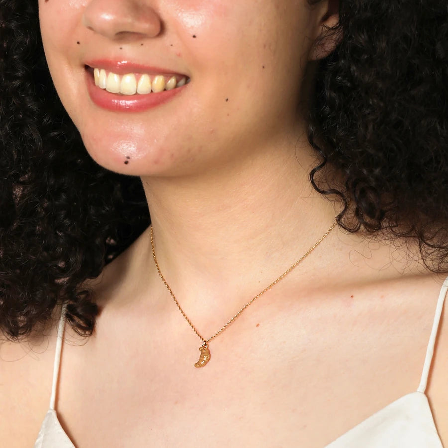 Gold necklace with a ceramic croissant pendant worn by a person with dark hair