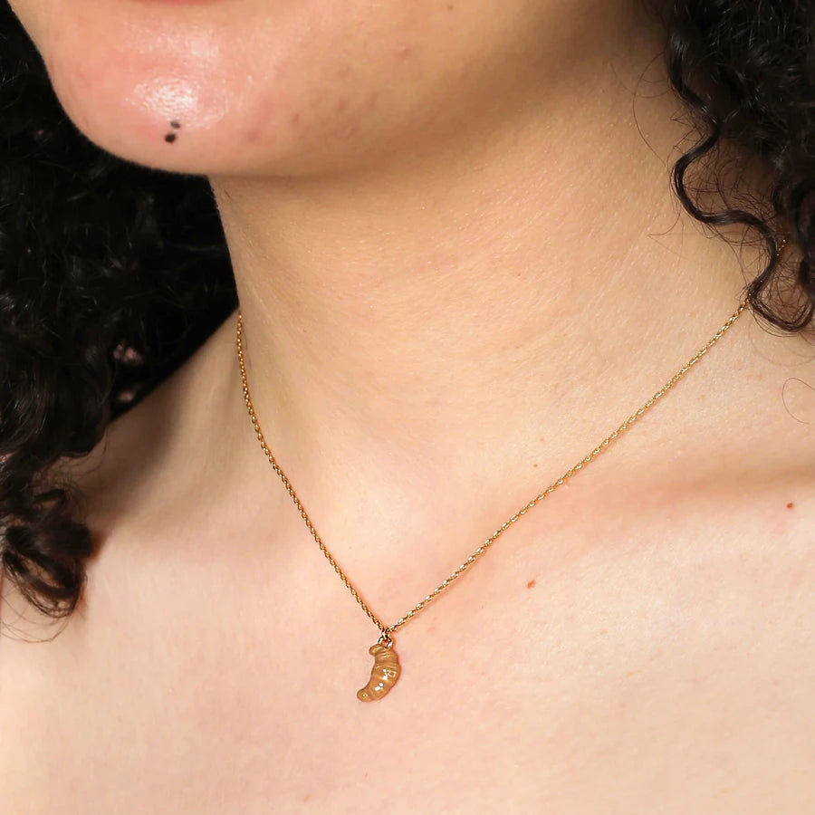 Gold necklace with a ceramic croissant pendant worn by a person with dark hair.