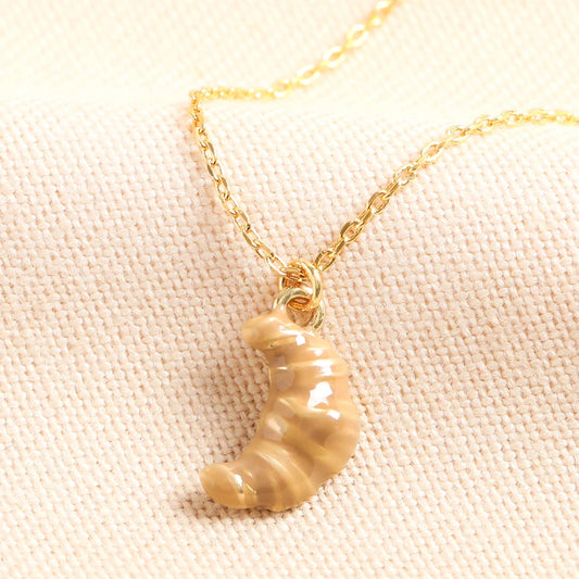 Gold necklace with a ceramic croissant pendant on a light pink fabric background